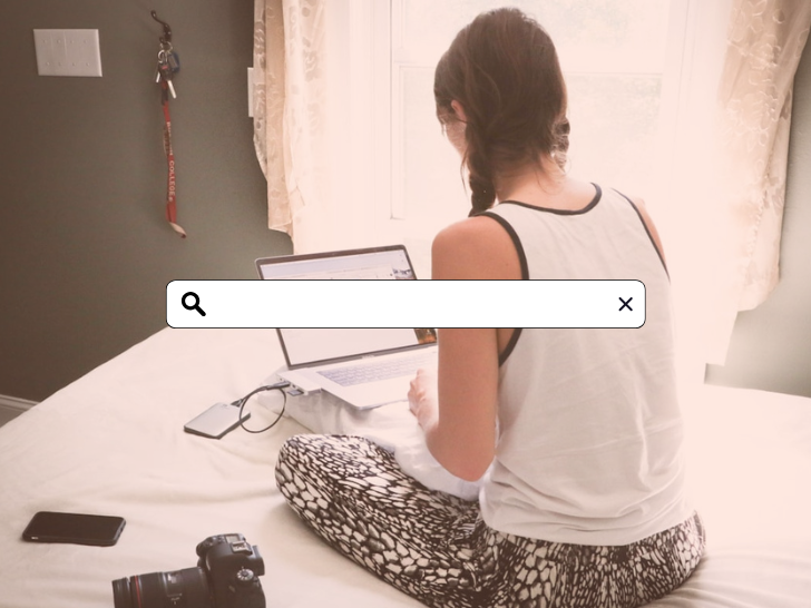 women sitting on bed working on laptop, facing away from viewer